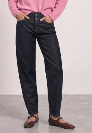 Personne portant un jean taille haute foncé avec double boutonnière, un pull rose et des chaussures Mary Jane marron, debout sur un sol gris.