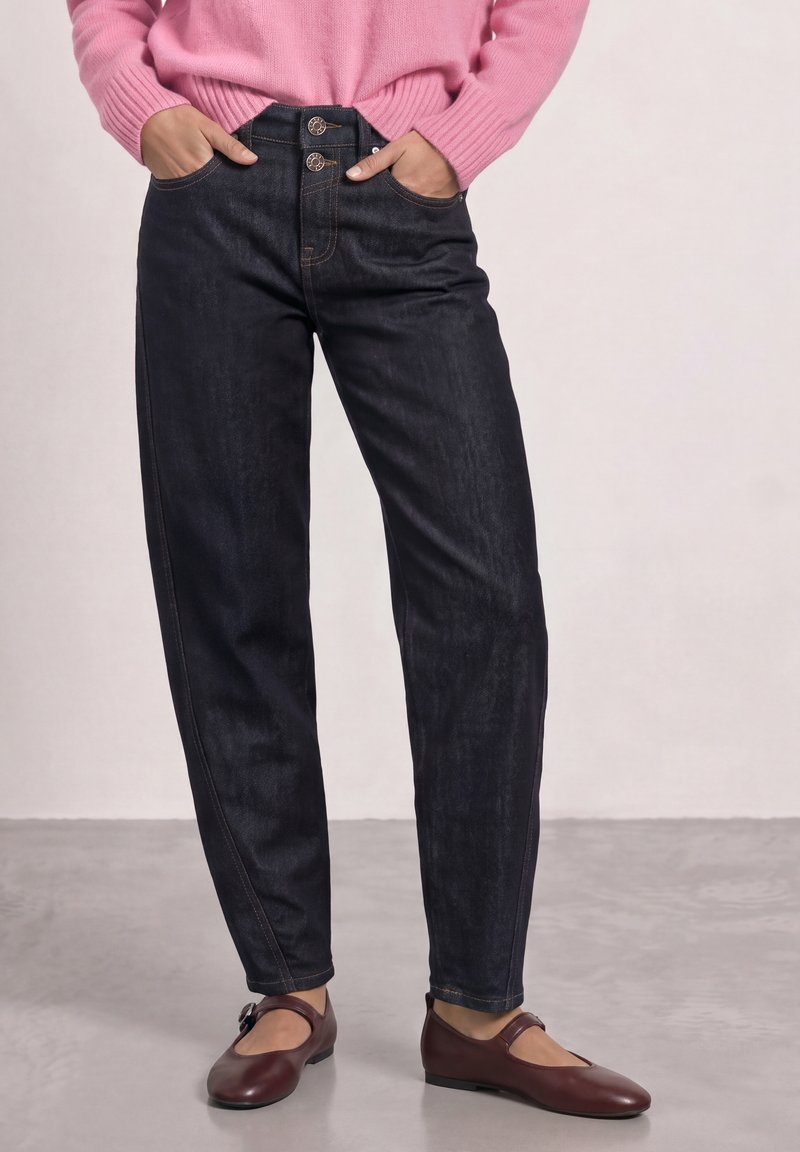 Personne portant un jean taille haute foncé avec double boutonnière, un pull rose et des chaussures Mary Jane marron, debout sur un sol gris.