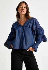 Denimbluse med volangærmer, dyb V-udskæring og frontknapper. Mørkeblå farve med en glat tekstur og sammensnørede manchetter.