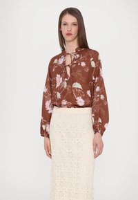 Bruine bloemenblouse met een keyhole-halslijn en lange mouwen, gecombineerd met een witte gehaakte rok met cirkelpatronen.