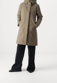 Lang hættejakke i lysebrun med lige snit, lynlås foran og sidelommer. Kombineret med vidde bukser i sort og sorte sko.