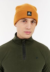 Beanie lavorato a maglia arancione con una toppa nera con il logo che presenta delle montagne; indossa un maglione in pile verde scuro a zip con un quarto di zip e tessuto testurizzato.