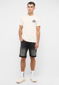 Beige short-sleeve t-shirt with navy graphic on left chest, paired with dark gray denim shorts and white sneakers with text accents.