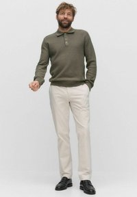 Olivgrön ribbad polo-sweater med tre knappar, ihop med gräddfärgade byxor och svarta loafers, mot en neutral bakgrund.