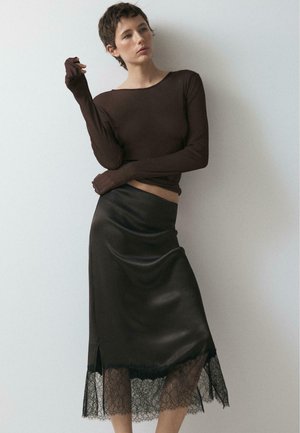 Žena s krátkými vlasy, která má na sobě těsné hnědé tričko s dlouhým rukávem a černou saténovou midi sukni s krajkovým lemováním, pózuje před jednoduchou bílou stěnou.