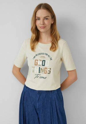 T-shirt crème à manches courtes avec un col rond, portant un texte imprimé de différentes couleurs disant "De bonnes choses sont à venir."