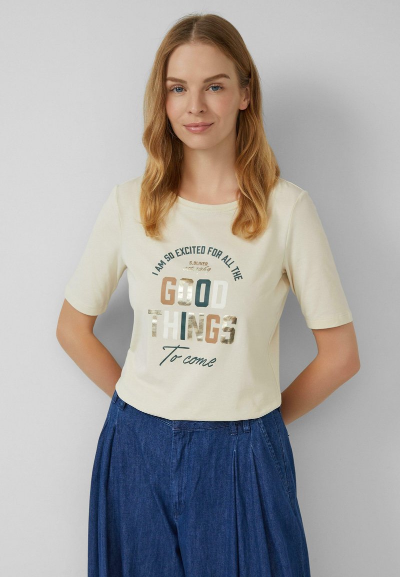 T-shirt crème à manches courtes avec un col rond, portant un texte imprimé de différentes couleurs disant "De bonnes choses sont à venir."