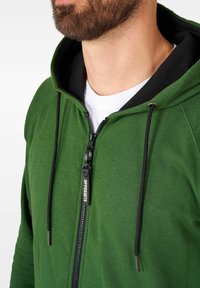 Chaqueta de capucha verde con forro interior negro, cordones y un tirador de cremallera con logo. Tejido liso con diseño ajustado y capucha.