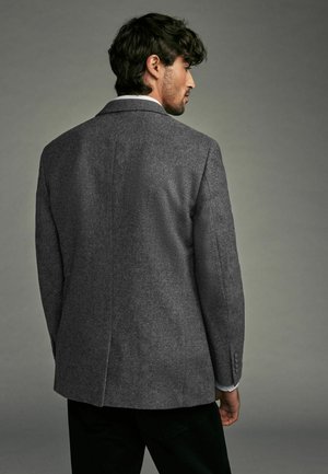 Grauer Fischgrat-Blazer mit strukturierten Schultern, Reverskragen und doppelten Schlitzen, kombiniert mit dunklen Hosen. Professionelles Aussehen.