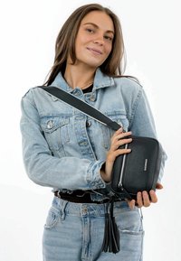 Borsa a tracolla in pelle nera con finitura testurizzata, zip argentata e dettaglio con nappina, tenuta da una persona che indossa una giacca di jeans.