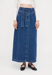 Denimmaxiskjørt med høy midje, knytbelte, to frontlommer og rett silhuett. Avsluttet med kontrastsøm og hemmet kant.