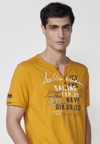 Geltonas medvilninis marškinėlis su apvaliu iškirpimu, trumpomis rankovėmis ir balta bei tamsiai mėlyna atspausdinta tekstu. Yra mygtuko detalė ir logotipas ant rankovės.