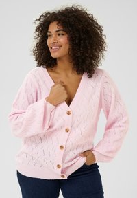 Světle růžový pletený cardigan s V-čelním výstřihem, zdobený zigzagovým vzorem a kulatými hnědými knoflíky, dlouhými rukávy a volným střihem.