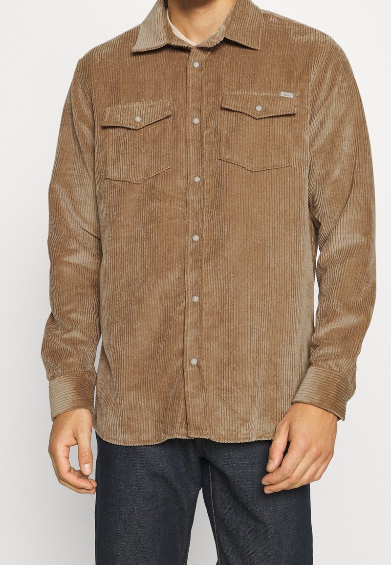 Homme portant une chemise boutonnée en velours côtelé marron clair avec deux poches à rabat sur la poitrine et un jean foncé, debout les mains détendues.