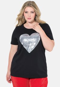 Černé tričko s krátkým rukávem, které má na přední straně velký srdcový design z velkých stříbrných flitrů. Tkanina je měkká s hladkým povrchem.