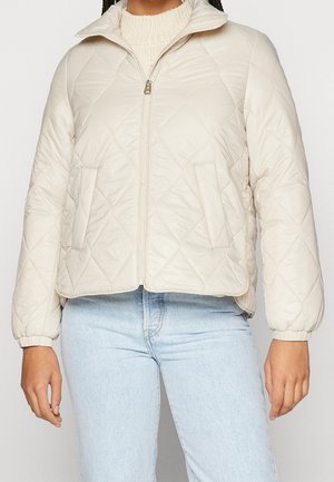 Personne portant une veste matelassée beige clair zippée par-dessus un pull en tricot crème et un jean bleu clair, debout les bras détendus.