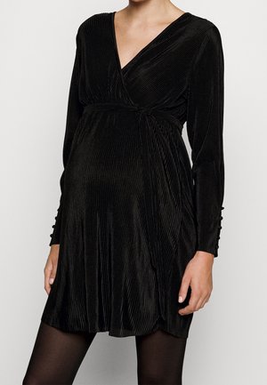 Femme portant une robe portefeuille en velours noir à manches longues, avec des plis verticaux et des détails de boutons sur les manches, debout devant un fond uni.