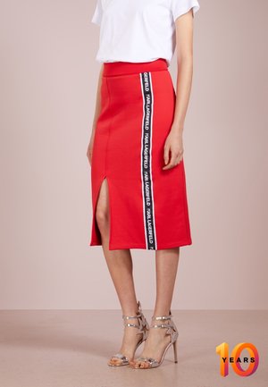 Femme portant une jupe midi rouge avec une fente sur le côté et une bande à logo noir et blanc, associée à des sandales à talons argentées et un haut blanc.