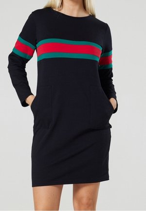 Vrouw draagt een zwarte jurk met lange mouwen met groene en rode strepen over de borst en mouwen, handen in de voorzakken.
