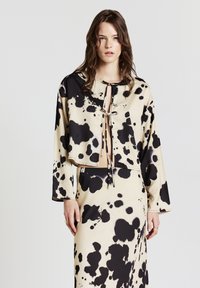 Donna che indossa un completo due pezzi astratto a macchie beige e nere con maniche lunghe e lacci davanti, su uno sfondo chiaro e semplice.