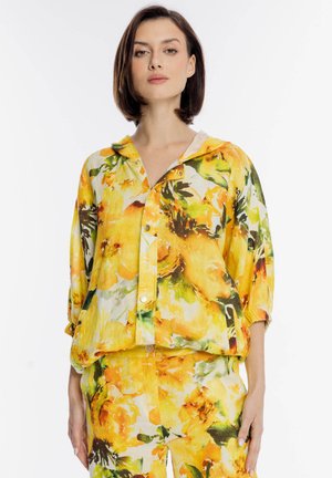 Kobieta ubrana w żółtą kurtkę w kwiatowy wzór z zamkiem błyskawicznym i zatrzaskami, w parze z pasującymi spodniami, stoi na białym tle.