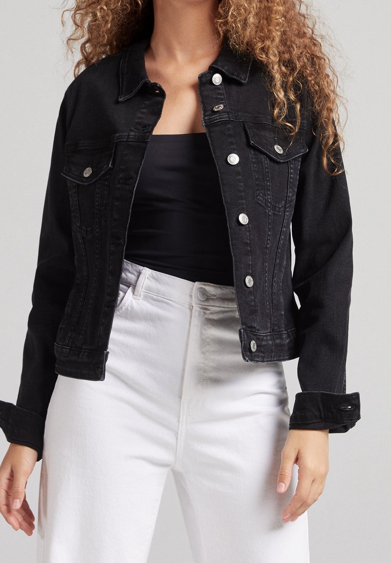 Black denim jacket with silver buttons, two chest pockets, and a fitted design. Worn over a black top and paired with white jeans.