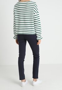 Henkilö seisoo selkä kameraan päin, päällä vihreä-valko-ravi takki, tummat kapeat housut ja valkoiset tennarit valkoista taustaa vasten.