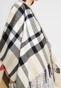 Femme portant un poncho en laine à carreaux beige, noir et blanc avec des franges, sur une tenue multicolore à carreaux, devant un fond uni.