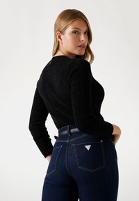 Maglione nero con trama, maniche lunghe, abbinato a jeans a vita alta blu scuro con un patch a forma di triangolo e dettagli dorati sulle tasche.