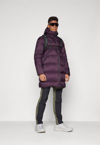 Paarse pufferjack met een hoge kraag, ritssluiting en zijzakken. Combinatie met zwarte en olijfgroene broek en lichtgrijze sneakers.