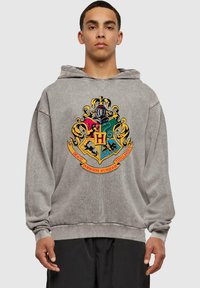 Sudadera con capucha gris que presenta un colorido escudo bordado de Hogwarts con varios símbolos, contorneado en amarillo y con una banda debajo.