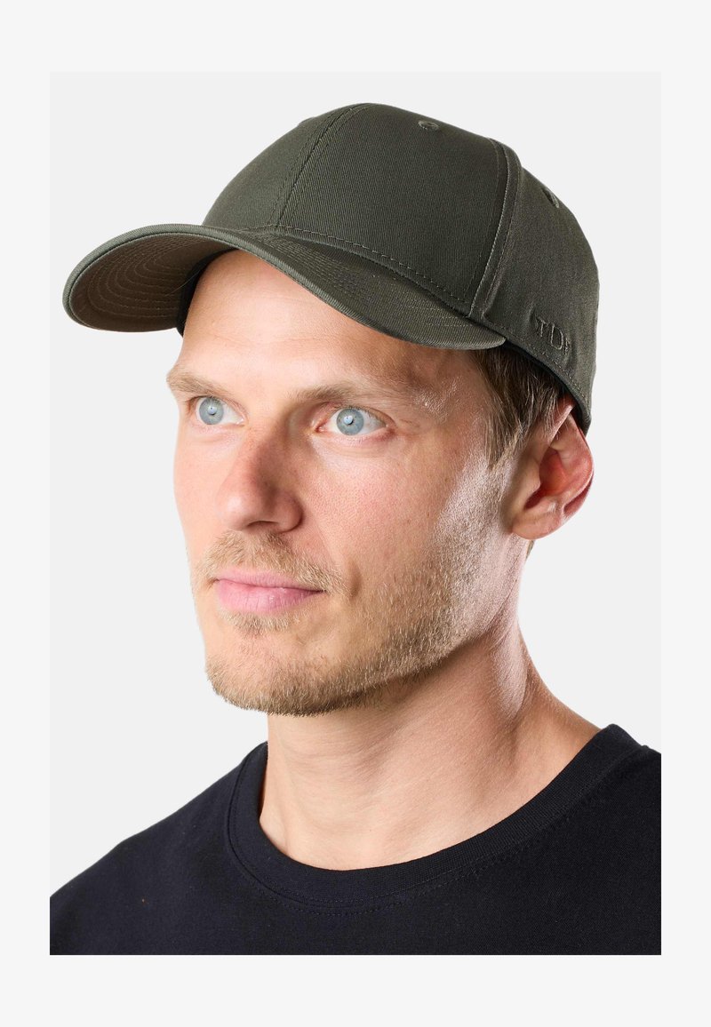 Joven con ojos azul claro que lleva una gorra de béisbol verde oliva y una camiseta negra de cuello redondo, mirando ligeramente hacia la derecha.