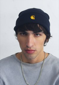 Berretto lavorato a maglia blu navy con un piccolo logo dorato. Il modello indossa una maglietta a maniche lunghe grigia chiara a coste e una collana in argento.