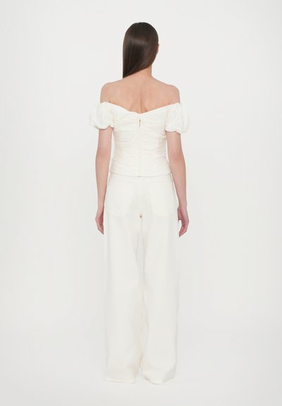 Off-the-shoulder, white top with puff sleeves and a ruched design, paired with wide-leg white trousers. Smooth fabric and zipper detail at back.