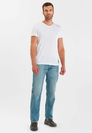 Man die recht vooruit staat, gekleed in een effen wit shirt met korte mouwen, lichtblauwe spijkerbroek en donkere casual schoenen, tegen een witte achtergrond.