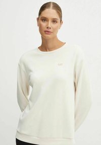 Felpa beige chiaro con collo rotondo, maniche lunghe, polsini a coste e un piccolo logo dorato sul petto. Tessuto dalla superficie liscia.