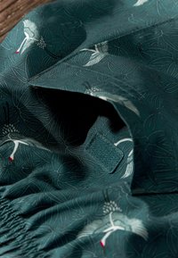 Tekstil dengan latar belakang teal tua dan pola halus burung bangau dan daun, dilengkapi dengan pengumpulan elastis dan tekstur yang halus.