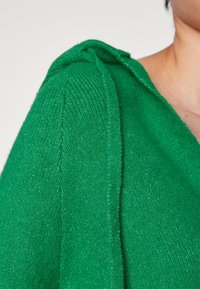 Gros plan d'une personne portant un vêtement tricoté vert vif avec un col en V et des détails de couture aux épaules, recadré au niveau du cou et du haut du bras.