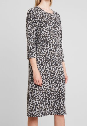 Vrouw draagt een knielange, lange mouw, jurk met luipaardprint en een ronde halslijn tegen een effen grijze achtergrond.