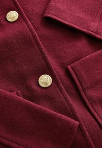 Burgundowy płaszcz z mieszanki wełny o klasycznym fasonie, z dwiema przednimi kieszeniami oraz wyraźnym złotym detalem w postaci guzika. Miękka tekstura, dobrze dopasowany krój.