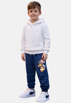 Young boy wearing a white hoodie, navy blue pants with cartoon puppy characters, and white sneakers with black accents, standing on a white background.