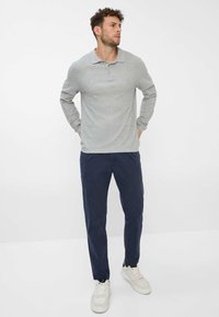 Helles graues Langarm-Polohemd mit Kragen, kombiniert mit marineblauen Hosen und weißen Sneakers, steht mit den Händen in den Taschen.