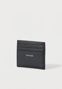 Μαύρη δερμάτινη θήκη καρτών με υφή στην επιφάνεια, έξι θήκες για κάρτες και ανάγλυφο λογότυπο Calvin Klein στο μπροστινό μέρος. Συμπαγής ορθογώνιο σχήμα.