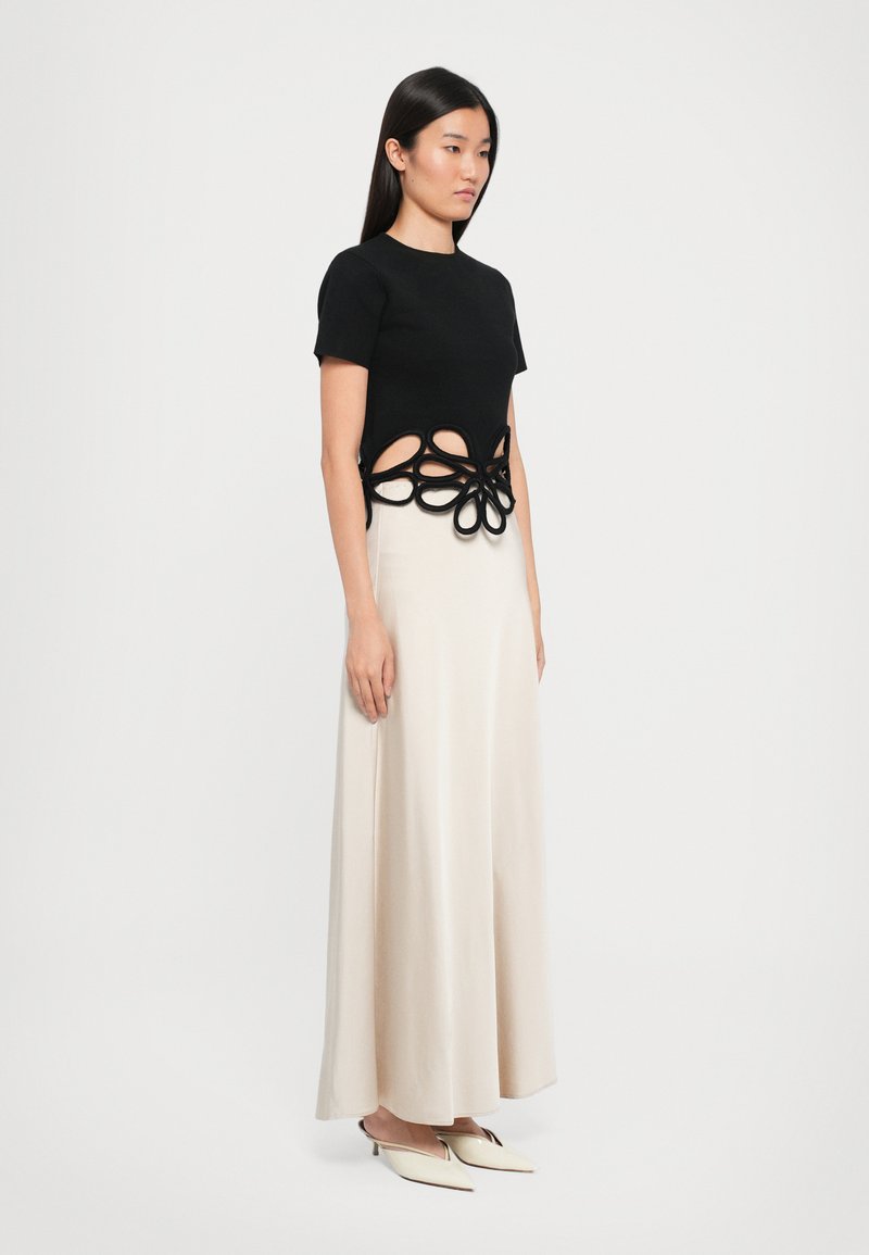 Haut noir à manches courtes avec un design floral découpé à la taille, associé à une jupe crème fluide et des chaussures blanches pointues. Textures lisses.
