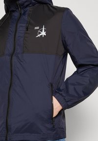 Giacca a vento militare con sezione superiore nera, dotata di zip frontale, polsini elasticizzati, tasche laterali e un logo stampato bianco sul petto.