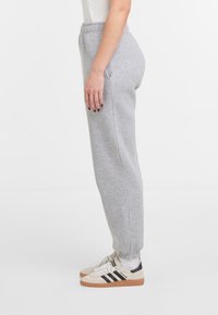 Personne portant un pantalon de survêtement gris clair et des baskets Adidas blanches avec des rayures noires, se tenant de profil sur un fond blanc.