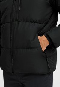 Sort pufferjakke med lynlås foran, sidelommer og elastiske manchetter, lavet af glat, isoleret materiale med mat finish.