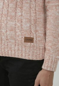 Maglione lavorato a maglia color rosa con motivo a trecce e orlo a coste, caratterizzato da un'etichetta in pelle marrone con la scritta "oxmo" nella parte inferiore.
