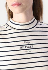 Kremo spalvos golfas su tamsiai mėlynomis horizontaliomis juostelėmis. Ant jo yra išspausdintas "Tommy HILFIGER" logotipas ir "Est. 1985" tamsiu tekstu. Pagamintas iš minkštos medvilnės.
