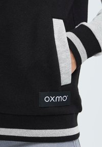 Sort sweatshirt med grå detaljer, med stribede manchetter og en sidelomme. Viser et 'OXMO' logo-tag i bunden.